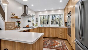 A modern kitchen with wooden cabinets, a stainless steel refrigerator, and a large window. - Maple Bluff Escape | A-Frame w/ Theater + Hot Tub (Stevens Point)
