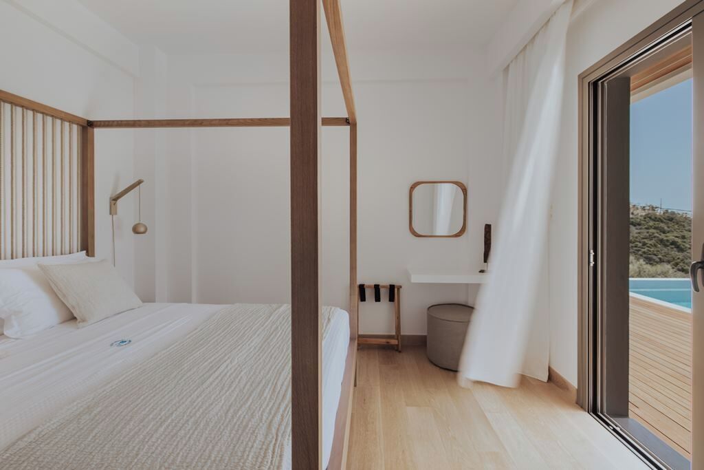 A modern bedroom with a bed, a wooden headboard, a bedside table, a mirror, and a view of a pool through a sliding door.