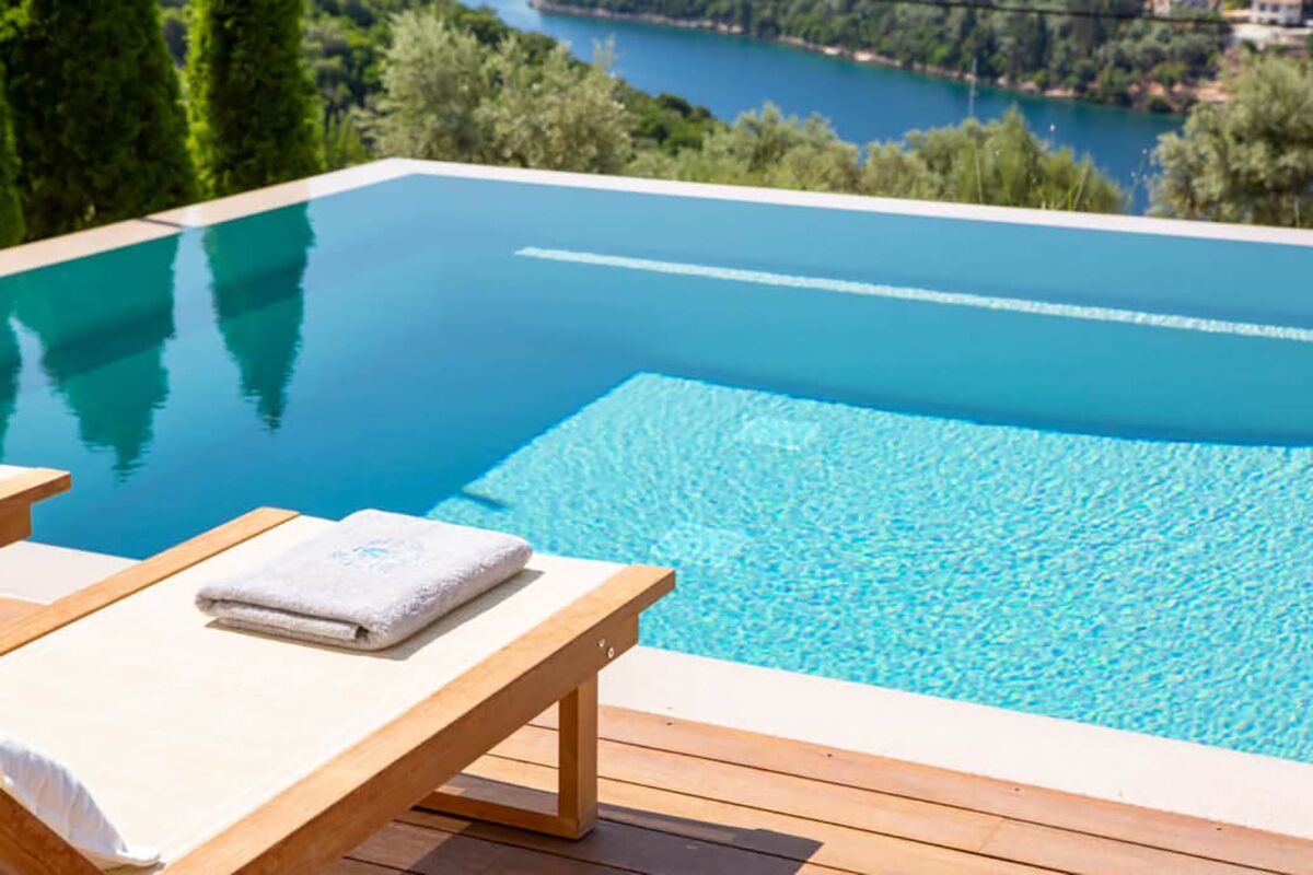Une magnifique piscine à débordement d’un bleu profond, dotée de chaises longues en bois et de serviettes, offrant une vue imprenable sur un lac pittoresque et une végétation luxuriante.