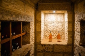 Um recanto com paredes de pedra, duas garrafas de vinho numa estante e uma estrutura de madeira para vinho à esquerda.