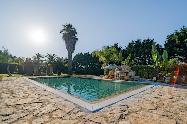 Piscina rectangular con borde de piedra, rodeada por un jardín con diversas plantas y árboles.