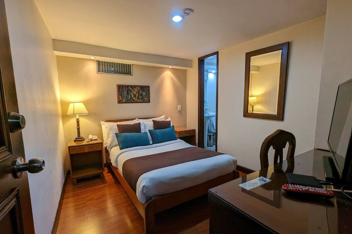 A hotel room with a bed, bedside table, lamp, and a desk with a mirror and remote control.