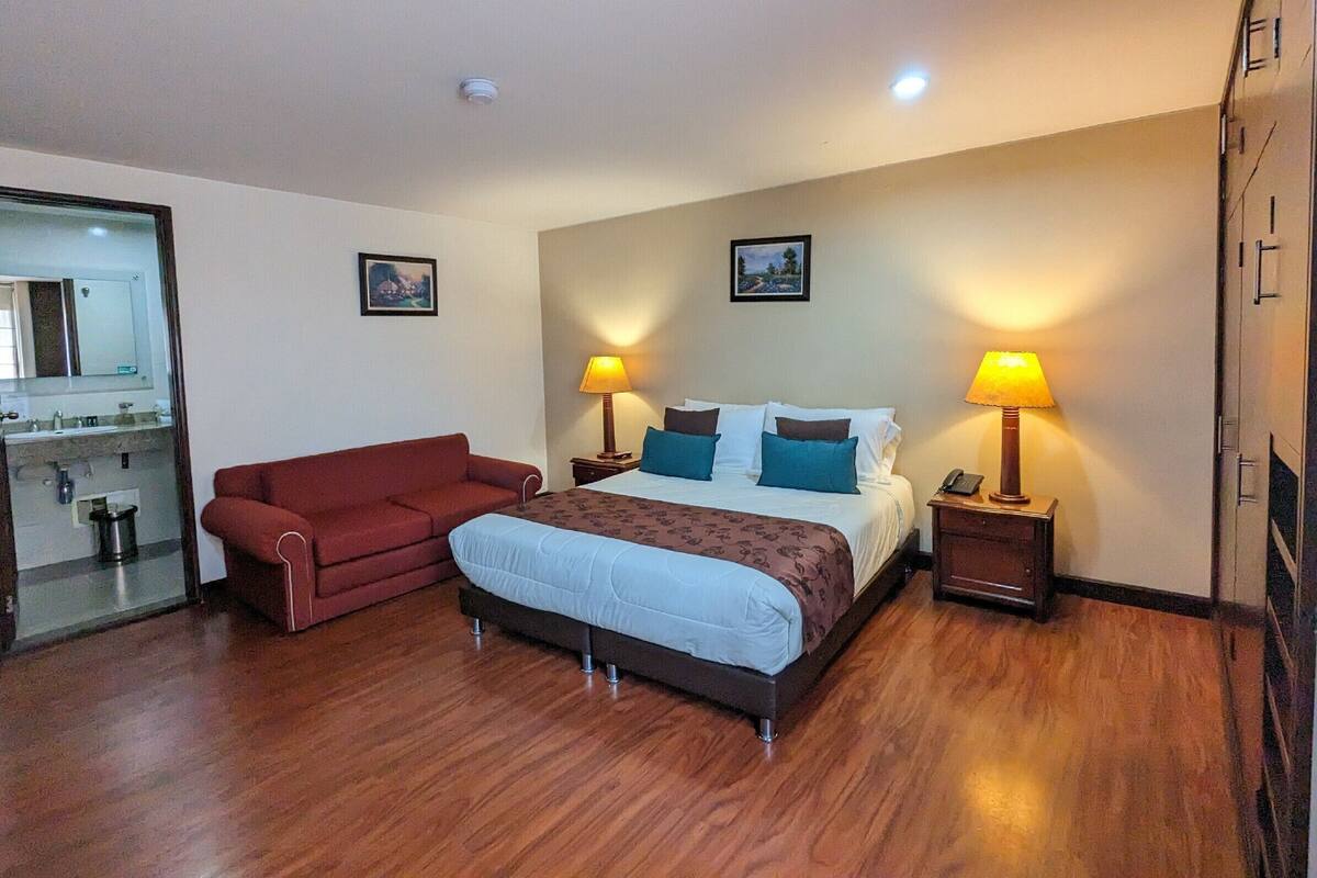 A hotel room with a bed, two bedside lamps, a red sofa, a wooden floor, a bathroom with a mirror, and two framed pictures on the wall.