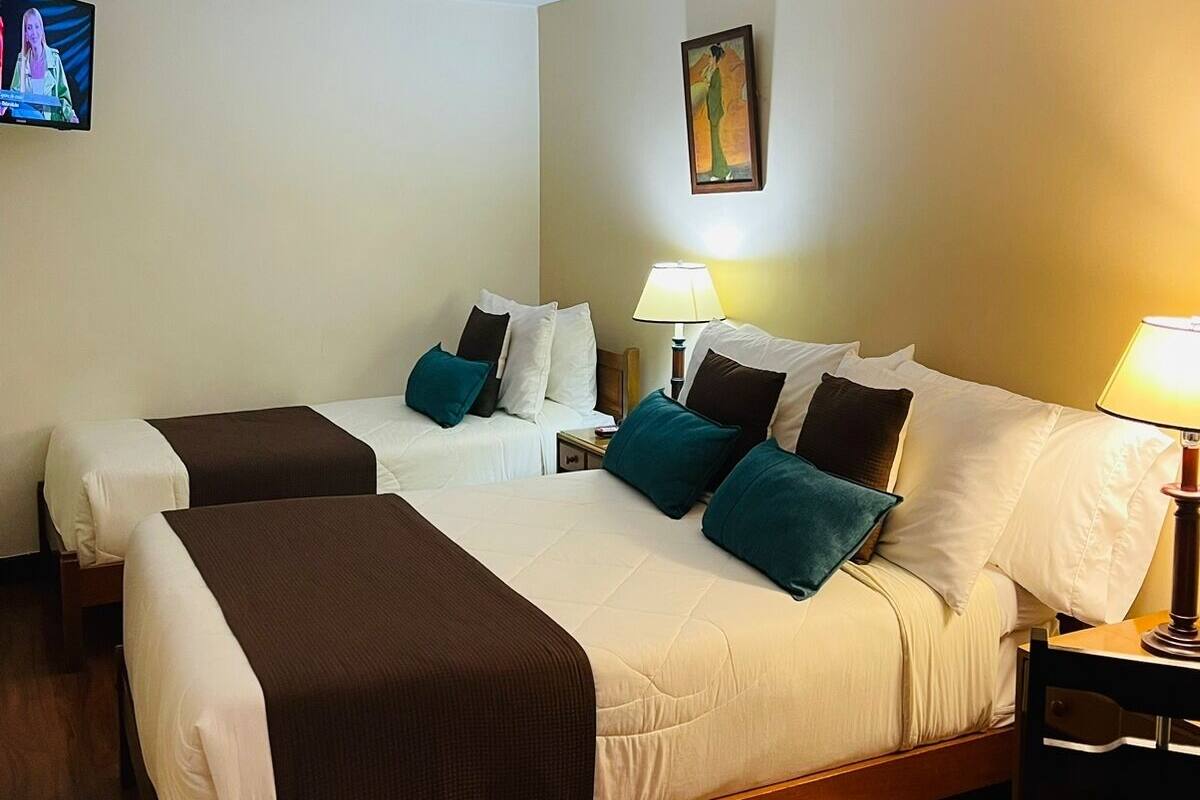 A hotel room with two beds, a TV on the wall, a painting, and a table with a lamp.