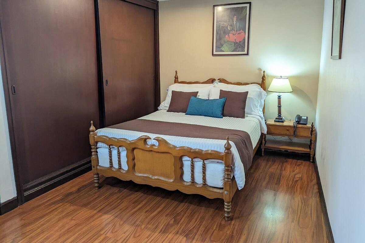 A neatly arranged bedroom with a wooden bed, a bedside table with a lamp, and a framed picture on the wall.