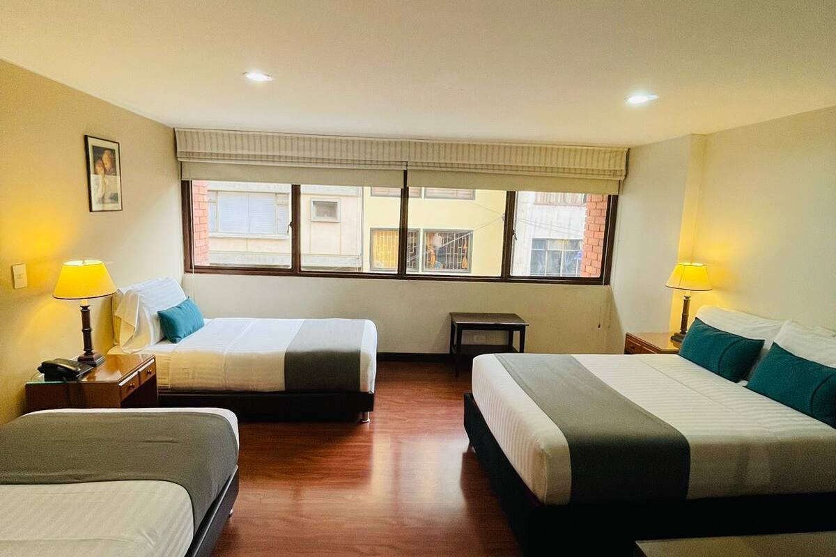 A hotel room with two beds, a window with blinds, a small table, and a lamp.