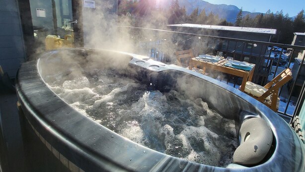 A hot tub with steam rising, surrounded by a wooden table and chairs, with a mountain view in the background. - Cozy Sunset | Amazing views | Private hottub (Revelstoke)