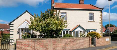 A two-story house with a brick wall in front, a red-tiled roof, and a small garden with yellow flowers.