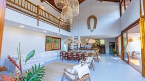 A spacious living area with a high ceiling, wooden staircase, and a dining area with chairs and tables.