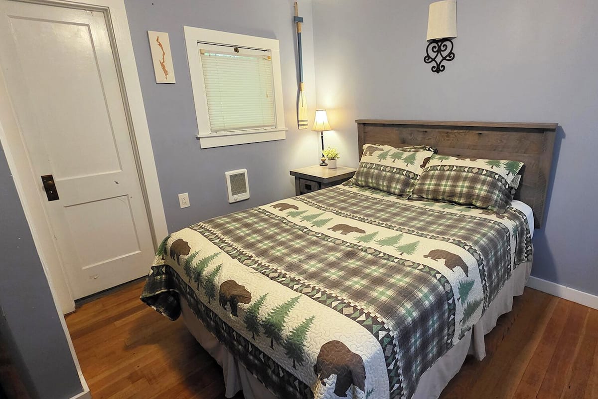 A bedroom with a bed, a nightstand, a lamp, a window with blinds, and a door.
