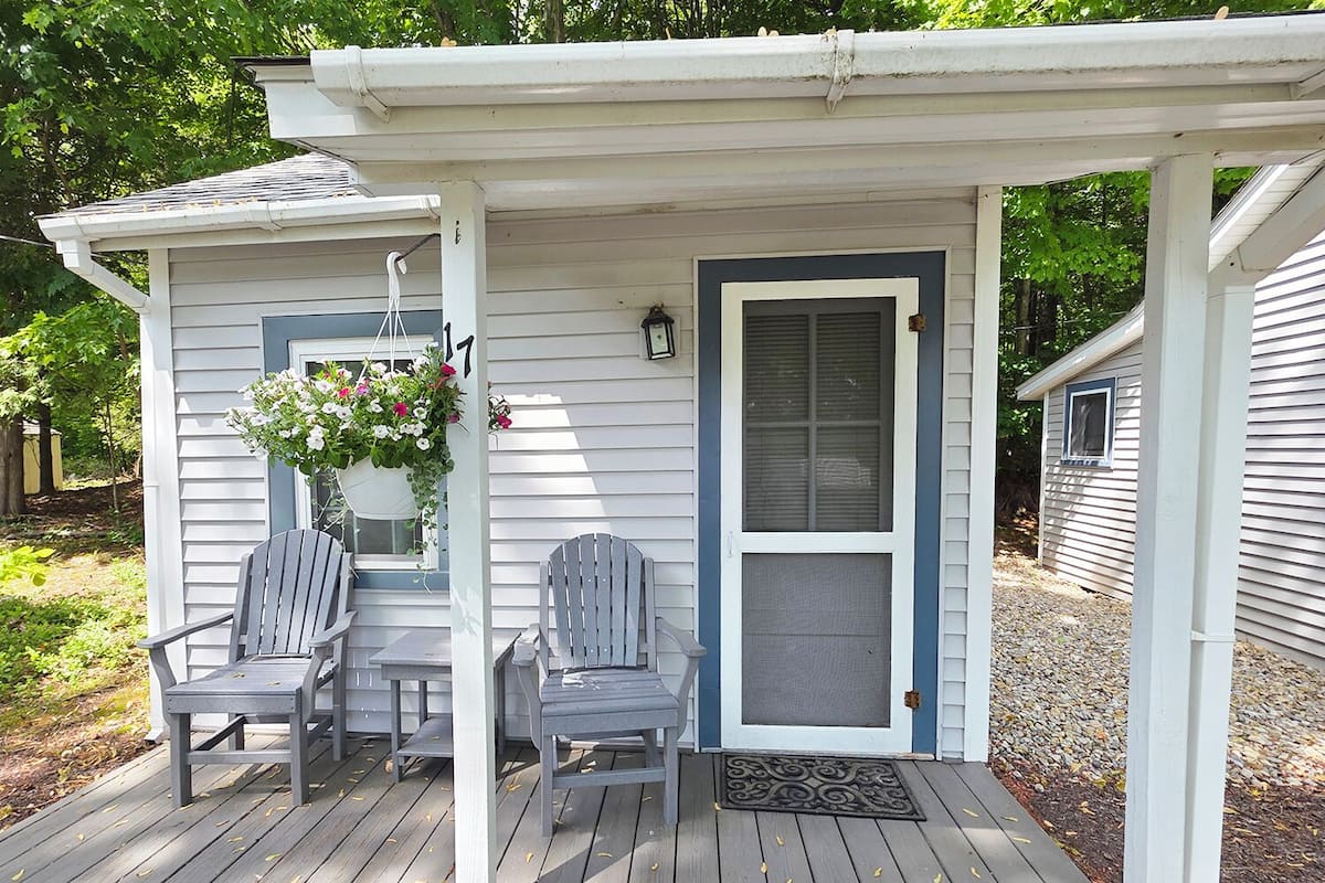 A covered porch with two Adirondack chairs, a hanging flower basket, and a door with a screen.
