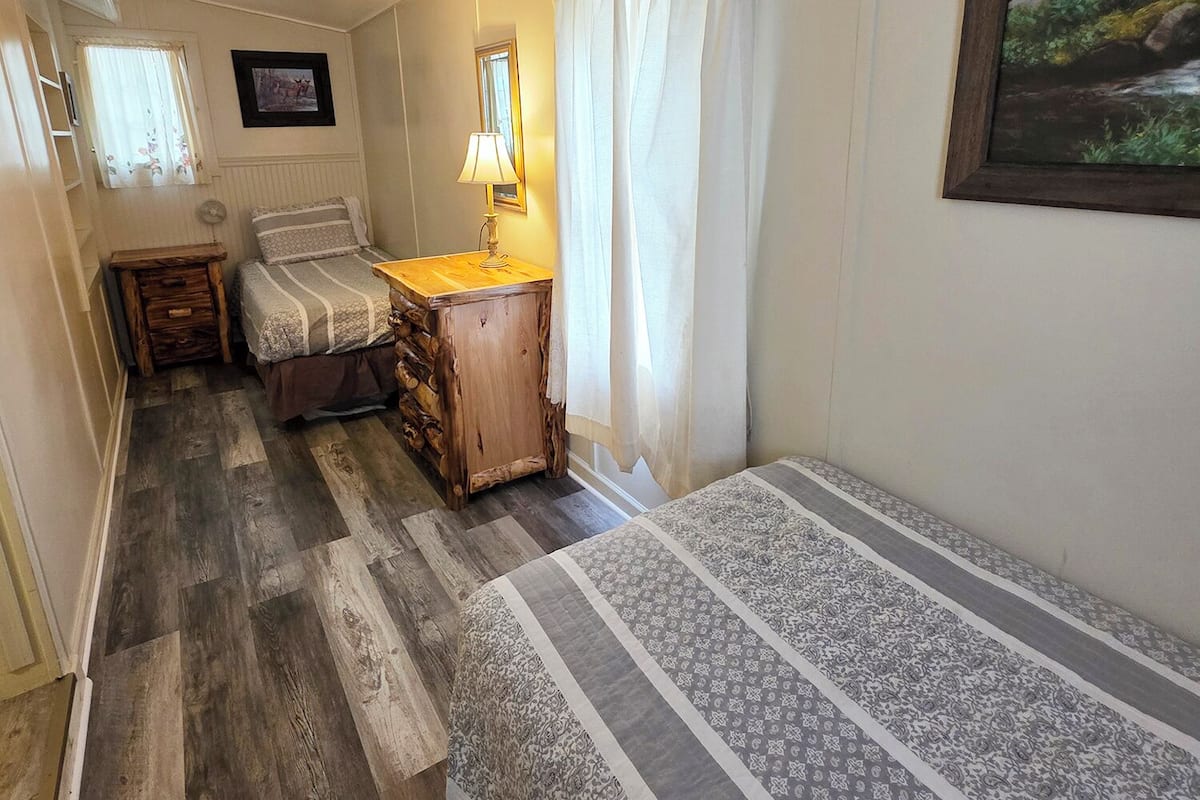 A narrow room with a bed, a wooden nightstand, a lamp, and a framed picture on the wall.