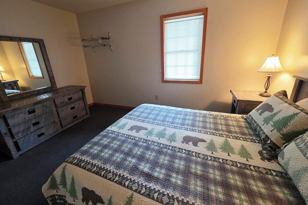 A bedroom with a bed, dresser, mirror, window, and bedside lamp.