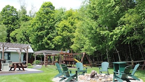 A backyard with a wooden swing set, picnic tables, and Adirondack chairs around a fire pit. - Adirondack Sunrise Motel and Cabins (Lake George)