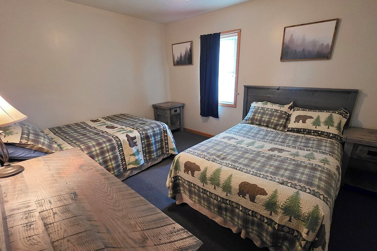 A room with two beds, a small table, a lamp, and a window with curtains.