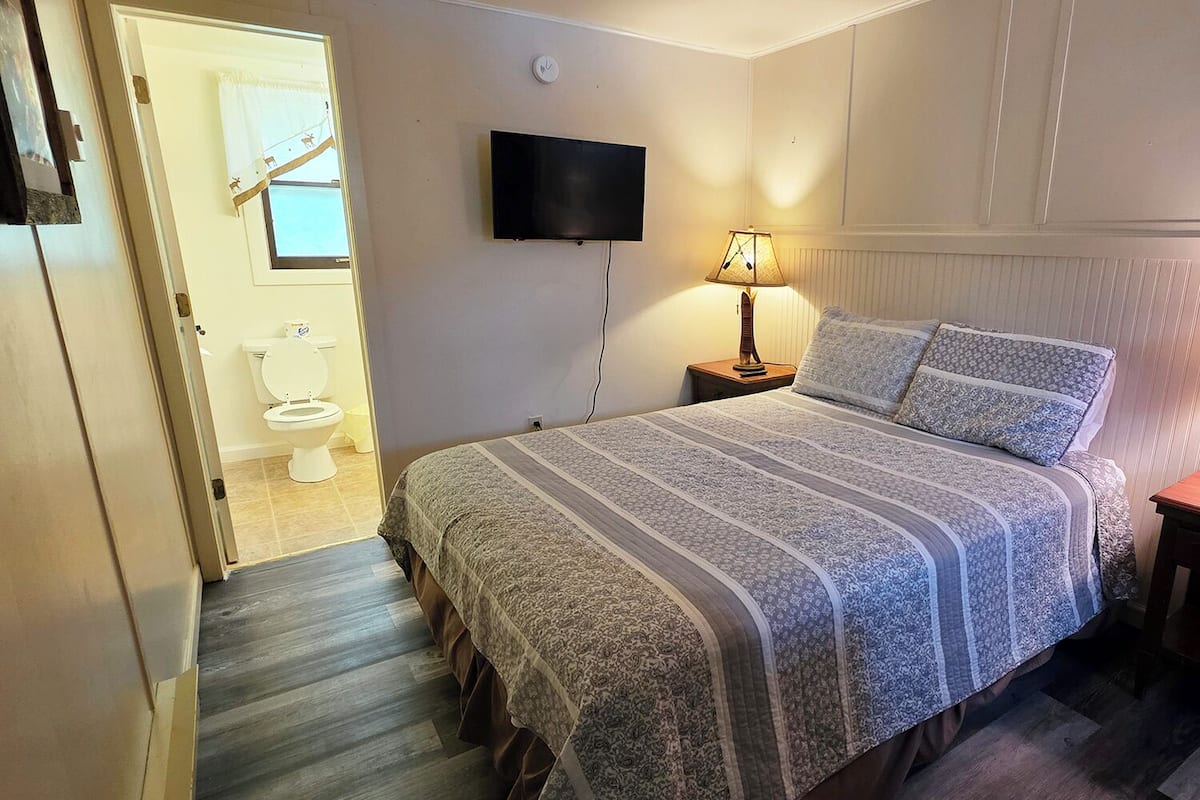 A bedroom with a bed, a television mounted on the wall, a bedside lamp, and a door leading to a bathroom.