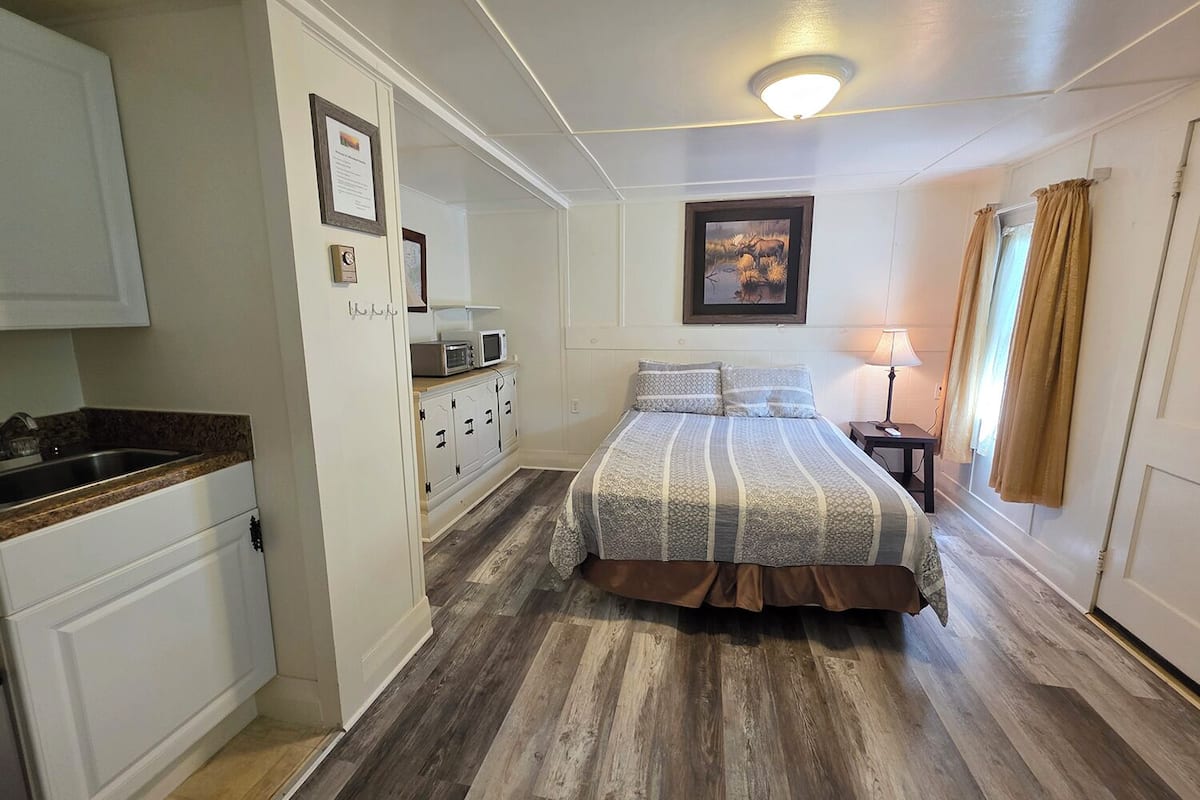 A bedroom with a bed, a bedside table, a lamp, a painting on the wall, and a kitchenette area with a microwave and cabinets.
