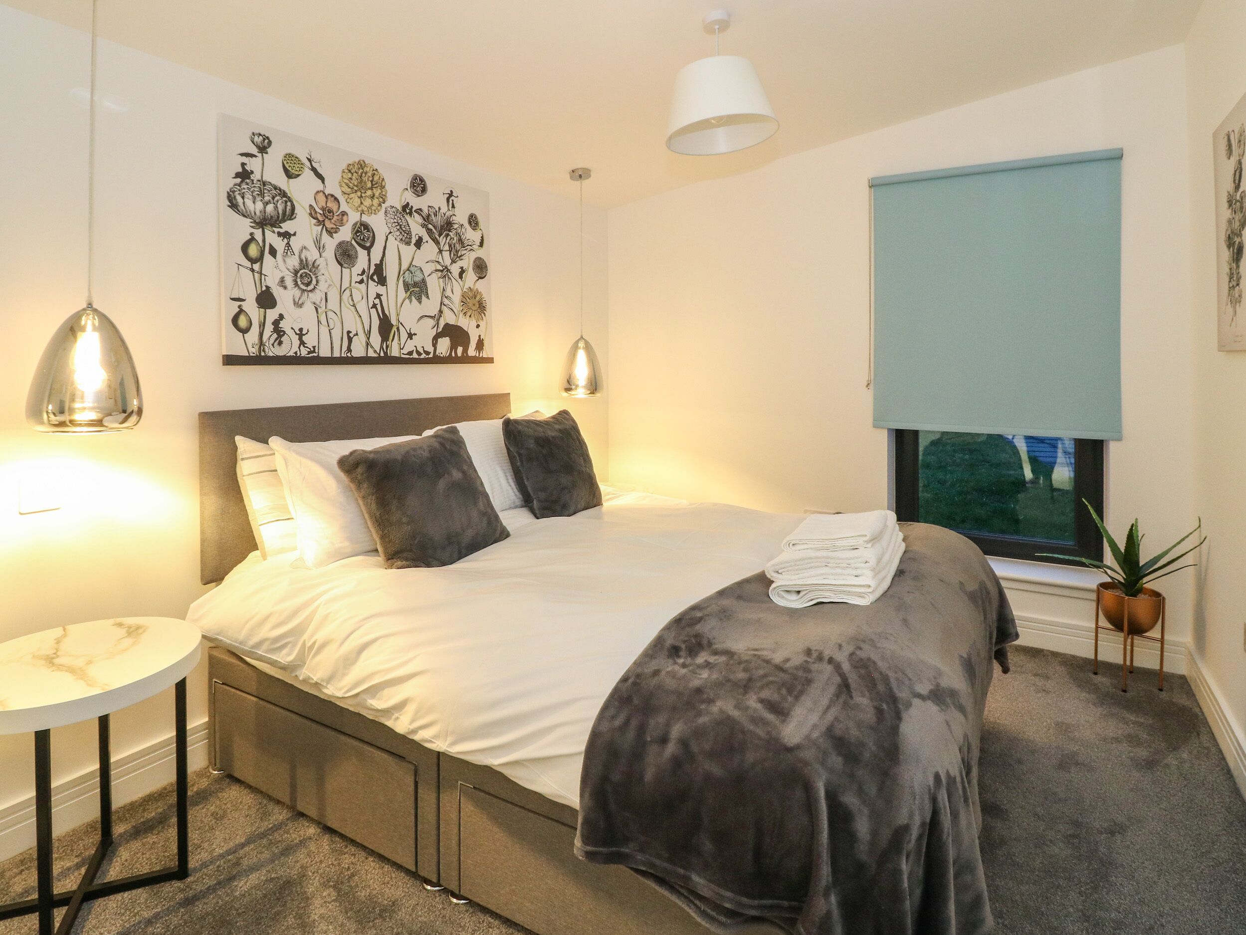 A neatly made bed with a grey blanket and pillows, a bedside table with a lamp, a wall art of flowers, a window with a view, and a small plant on the windowsill.