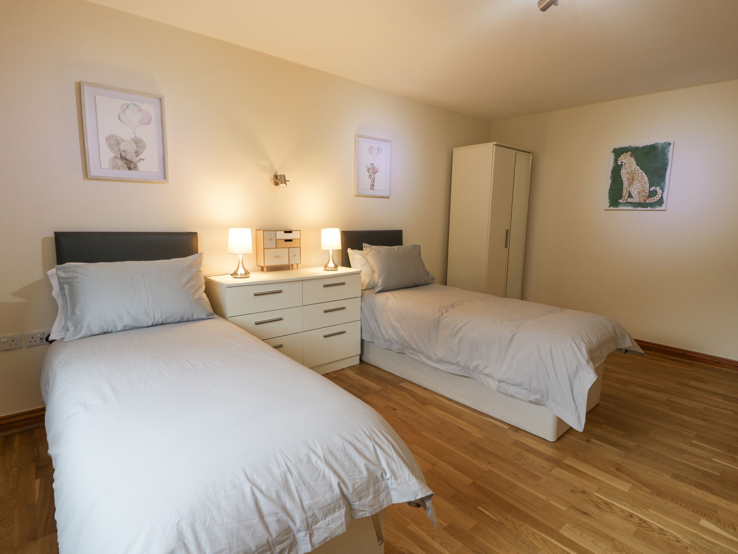 A hotel room with two single beds, a nightstand with lamps, a wardrobe, and artwork on the walls.