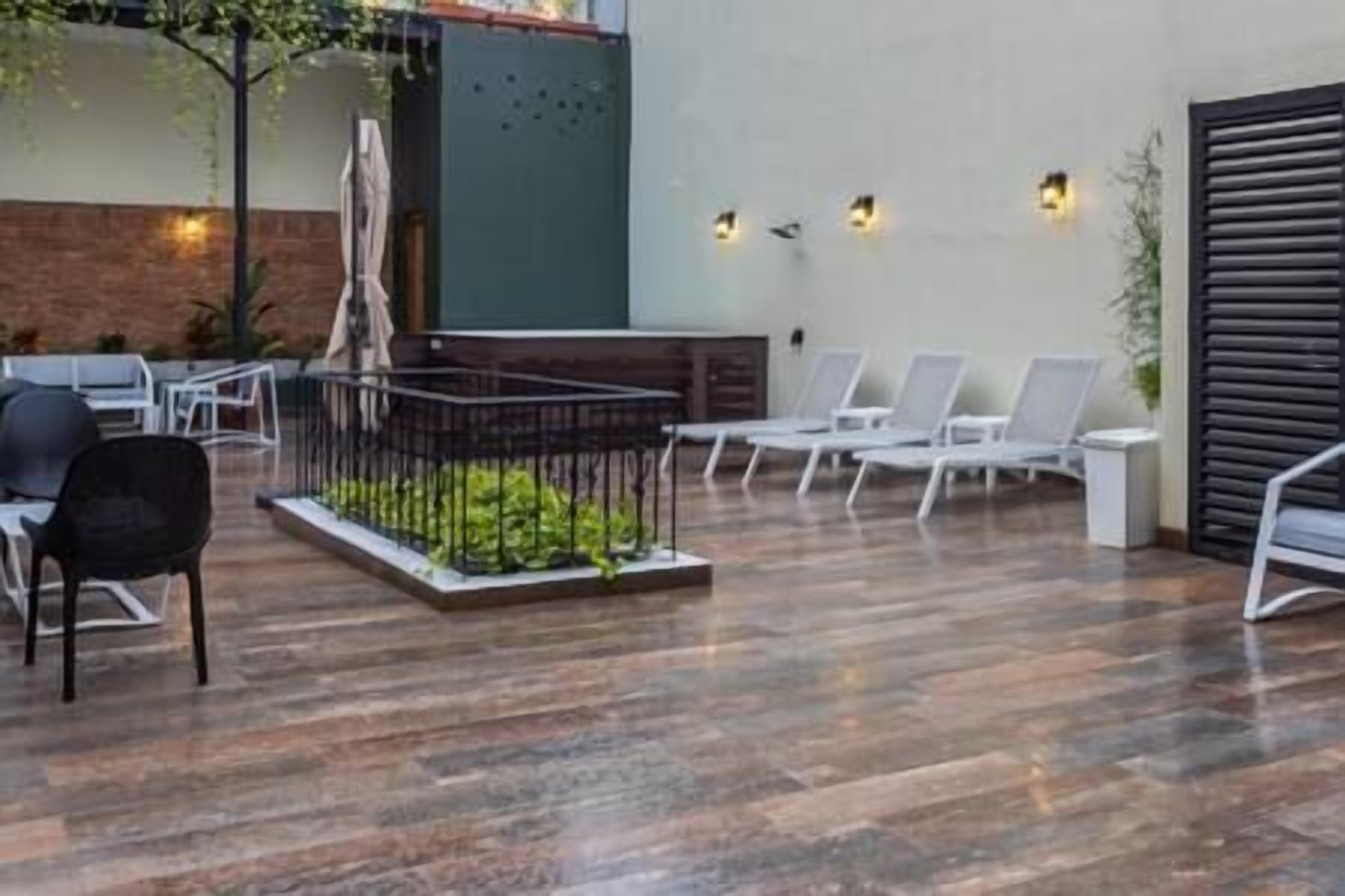 Un espacio exterior moderno con suelo de madera, sillones blancos y un pequeño jardín con valla metálica.