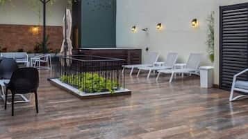 Un espacio exterior moderno con suelo de madera, sillones blancos y un pequeño jardín con valla metálica.