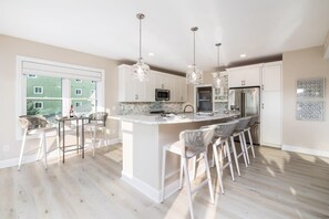 A modern kitchen with white cabinetry, a central island with bar stools, pendant lighting, and a view of a green building outside.