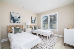 A bedroom with two beds, a window with blinds, and artwork on the walls.