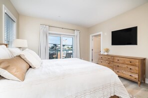 A bedroom with a large bed, a wooden dresser, a flat-screen TV, and a balcony with a view of buildings.