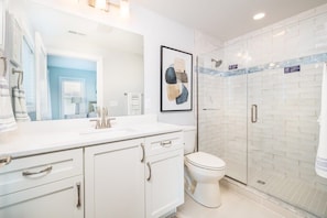 A modern bathroom with a white vanity, a large mirror, a framed abstract artwork, a glass shower enclosure, and a toilet.