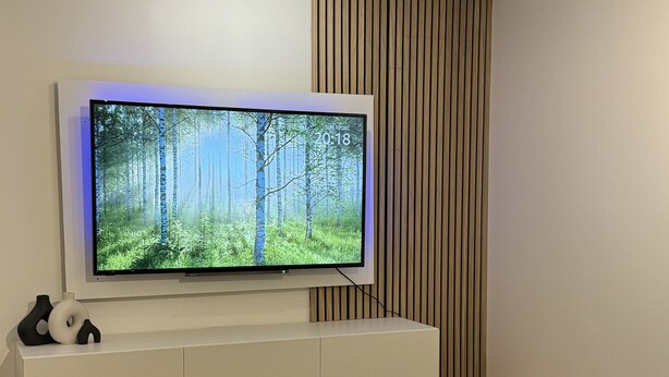 A modern living room with a flat-screen TV mounted on a white cabinet, a glass coffee table, a dark green sofa, and a wooden slat wall panel. - Cosy 1bed in Wembley  (Wembley)