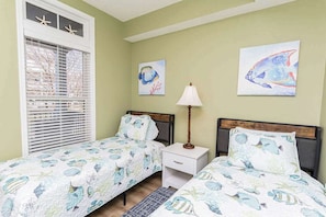 A bedroom with two beds, a window with blinds, a nightstand with a lamp, and two wall art pieces.