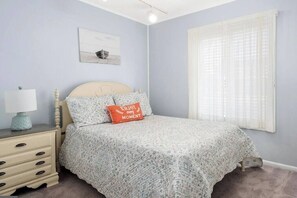 Un dormitorio con cama, mesita de noche, lámpara, ventana con cortinas traslúcidas y un cuadro colgado en la pared.