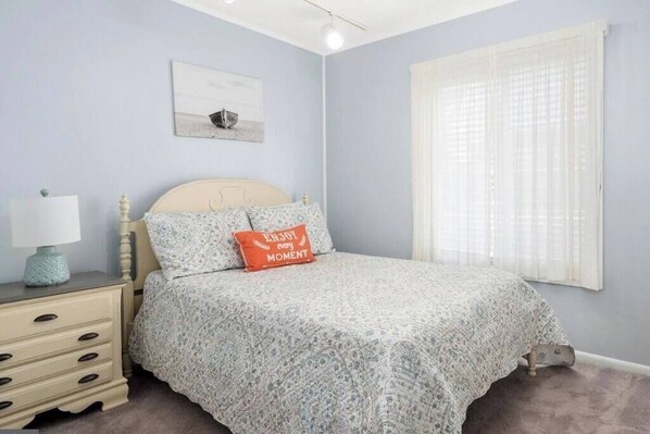 A bedroom with a bed, a nightstand, a lamp, a window with sheer curtains, and a wall-mounted picture.