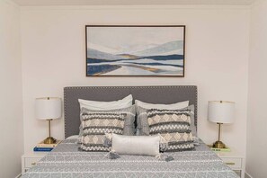 A bedroom with a bed, two bedside lamps, a framed picture above the bed, and a nightstand with a book.