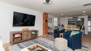 Un salon moderne doté d’un téléviseur à écran plat, d’un mur décoratif en briques, d’un coin repas avec une cuisine en arrière-plan et d’un canapé bleu agrémenté d’un coussin sur lequel est inscrit le mot « bienvenue ».