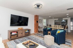 Ein modernes Wohnzimmer mit Flachbildfernseher, einer Ziegelfassade, einem Essbereich und einer dahinterliegenden Küche, sowie einem blauen Sofa mit Kissen, das den Schriftzug 'welcome' trägt.