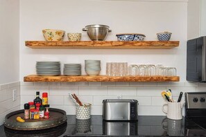A kitchen with wooden shelves holding bowls and plates, a round wooden table with condiments, a toaster, and a kettle.
