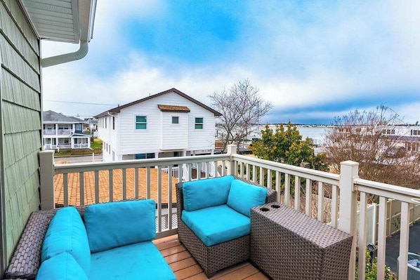 A balcony with wicker furniture and a view of a white house across the water.