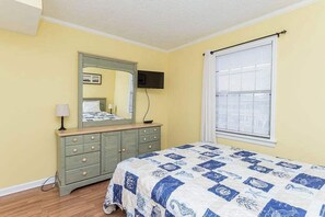 Un dormitorio con cama, cómoda, espejo, televisor y ventana con persianas.