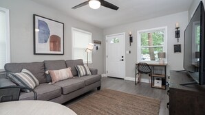 Ein Wohnzimmer mit grauem Sofa, Deckenventilator, Fernseher und einer Tür mit Fenster.