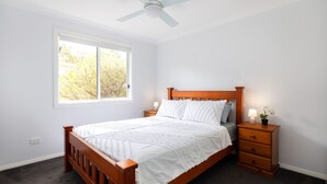 A bedroom with a wooden bed, a nightstand with a lamp, a window with a view of trees, and a ceiling fan. - Tallwood By The Sea in Mollymook (Mollymook)