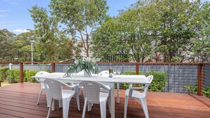 An outdoor dining area with a wooden deck, white plastic chairs, and a table set with a bottle and glasses. - Tallwood By The Sea in Mollymook (Mollymook)