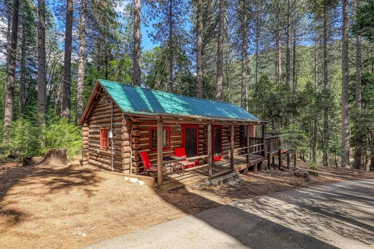 Uma cabana de tronco com telhado verde, varanda com cadeiras vermelhas e um caminho de terra que a leva.