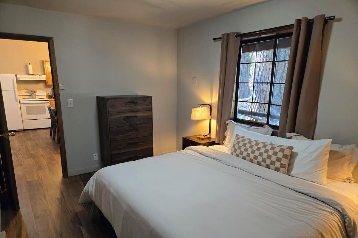 Quarto com cama, mesa de cabeceira, luminária, cômoda e janela com cortinas.