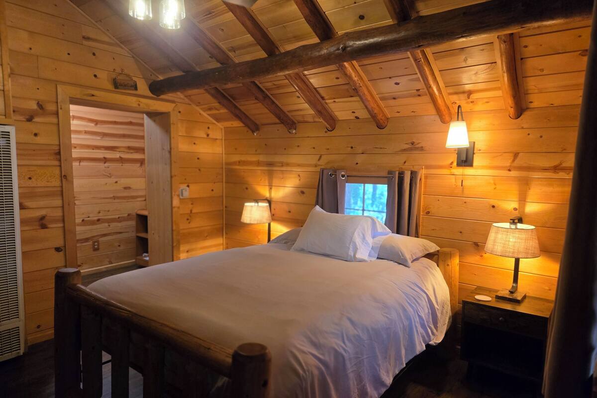 Quarto de cabana de madeira com cama, duas luminárias de apoio, janela com cortinas e teto de madeira.