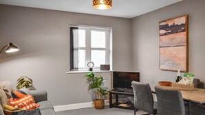 A modern living room with a grey sofa, a dining table with chairs, a TV, a window with curtains, and a painting on the wall. - The Gainsborough Mews (Gainsborough)