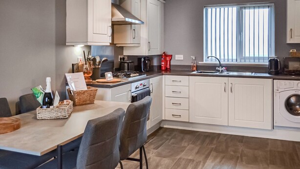 A modern kitchen with white cabinets, a dining area with a table and chairs, a built-in oven, and a window with blinds. - The Gainsborough Mews (Gainsborough)