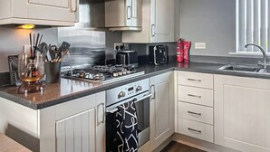 A modern kitchen with white cabinets, a stainless steel range hood, a gas stove, a coffee maker, and a window with blinds. - The Gainsborough Mews (Gainsborough)