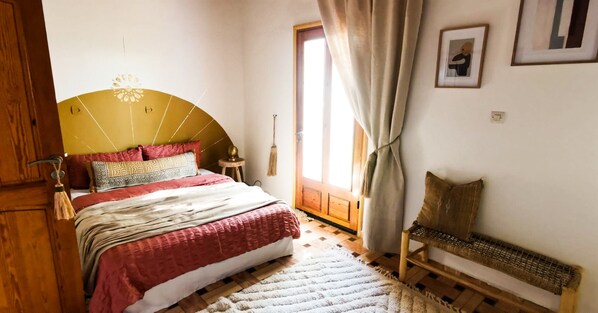 Ein Schlafzimmer mit einem Bett, einem Holzhocker, einem Fenster mit Vorhängen und einer Tür.