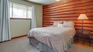 Een slaapkamer met een houten blokkenwand, een bed met een gebloemd dekbed, een nachtkastje met een lamp, en een raam met gordijnen.